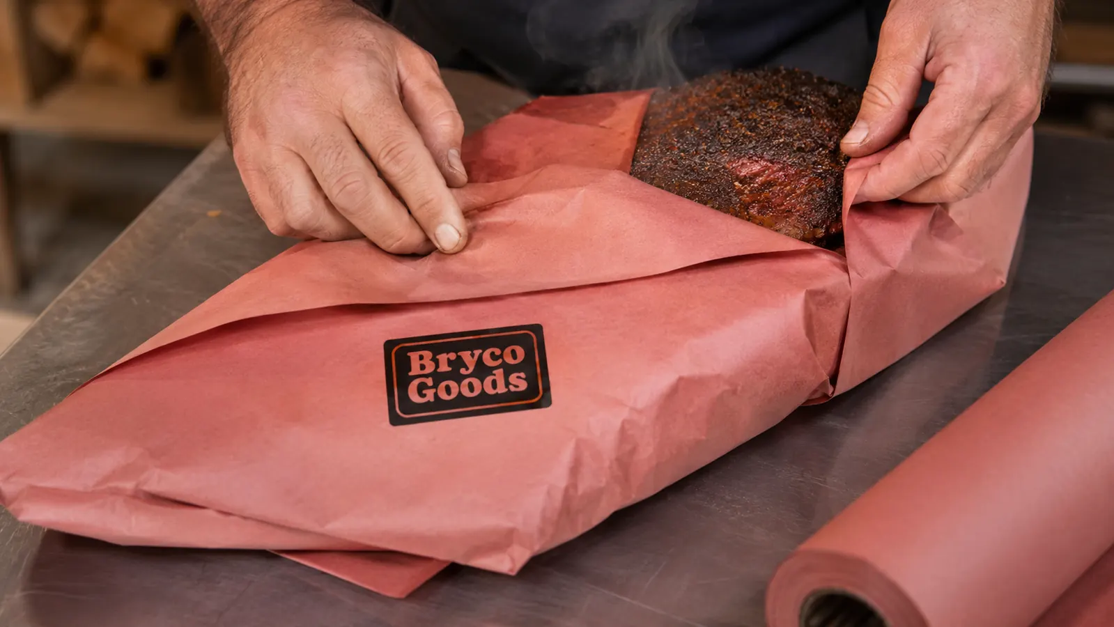 Weathered hands folding pink butcher paper tightly around a brisket on a stainless steel prep table