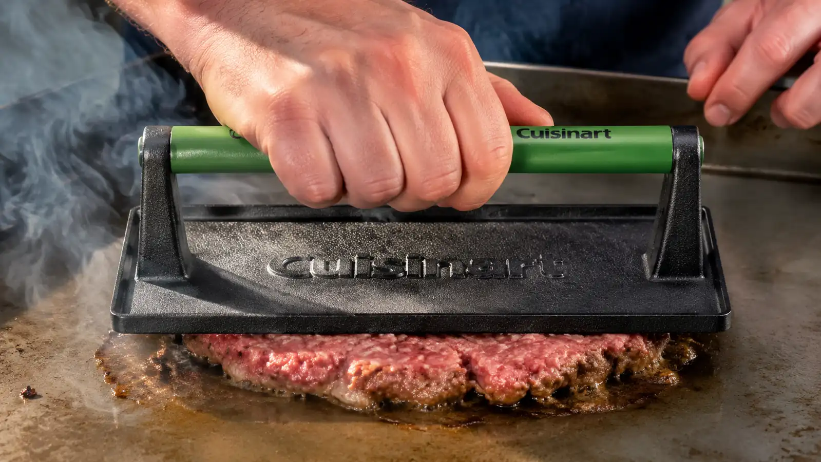 Hand pressing a wooden-handled cast iron press down onto a beef ball on a hot griddle, smoke rising off the crust
