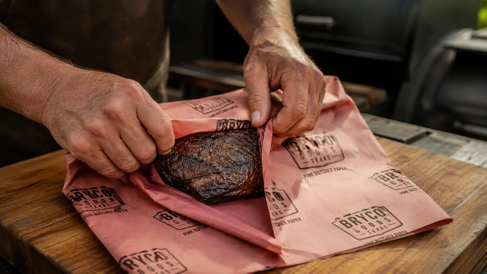 Hands wrapping a smoked brisket in pink butcher paper on a wooden cutting board next to a smoker