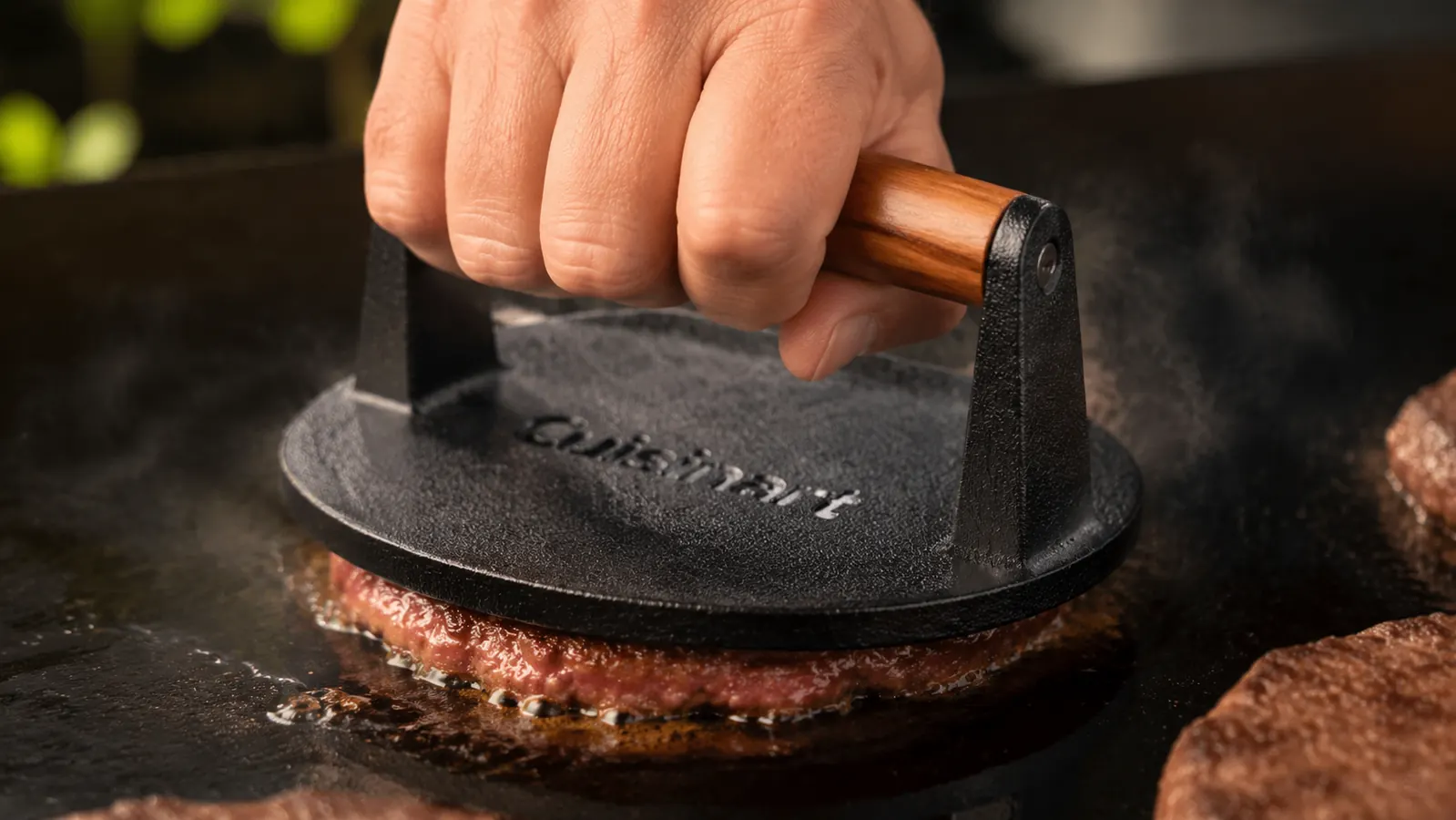 Close-up of a hand pressing a raw burger patty flat with a cast iron press on a hot griddle, creating a dark seared crust