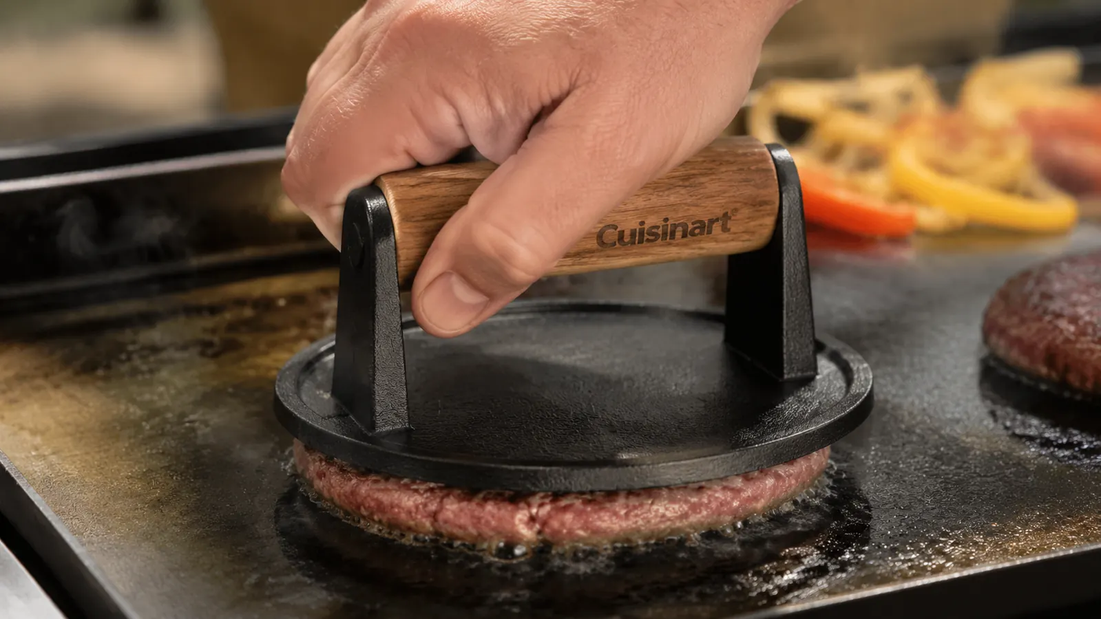 Hand pressing a burger patty down on a flat top griddle with the cast iron press, grease pooling at the edges