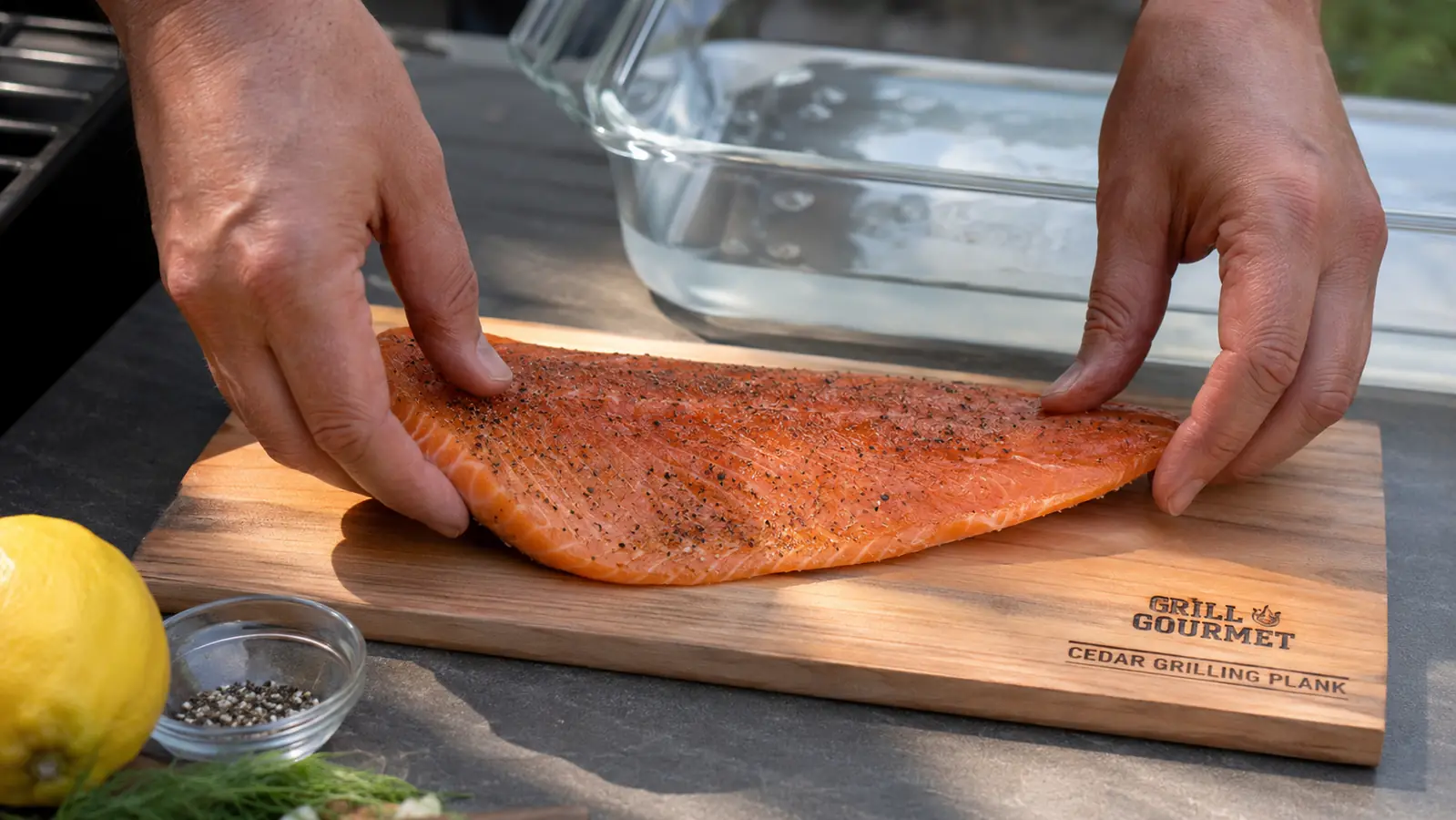 Hand placing a raw salmon fillet onto a soaked cedar plank next to a tub of water on a patio table
