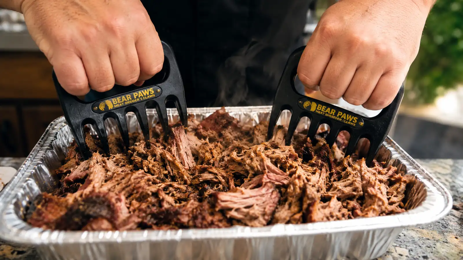 Hands using red-handled meat shredding claws to pull apart pork shoulder in a foil pan